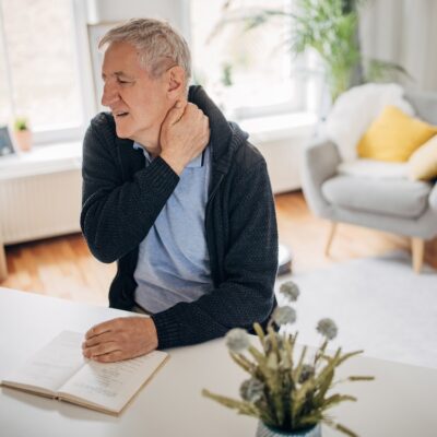 One man, old man having a neckache problems while sitting at home and reading a book.